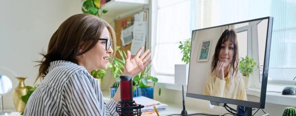 A woman seated at a desktop computer chats with an online student in a live video call.