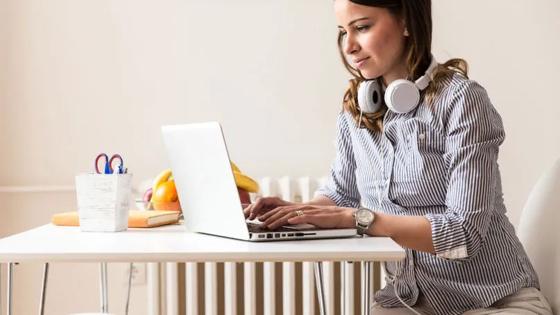 Woman with headphones around her neck types on a laptop seated at a kitchen table.