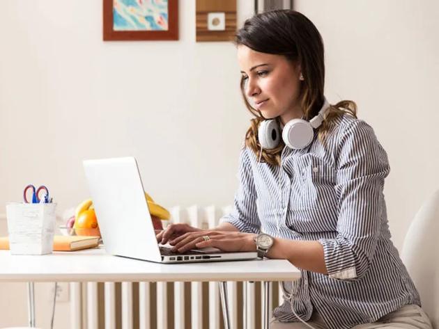 Woman with headphones around her neck types on a laptop seated at a kitchen table.