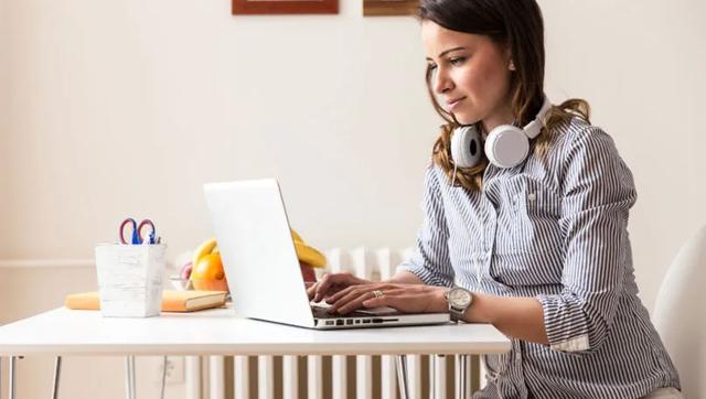 Woman with headphones around her neck types on a laptop seated at a kitchen table.