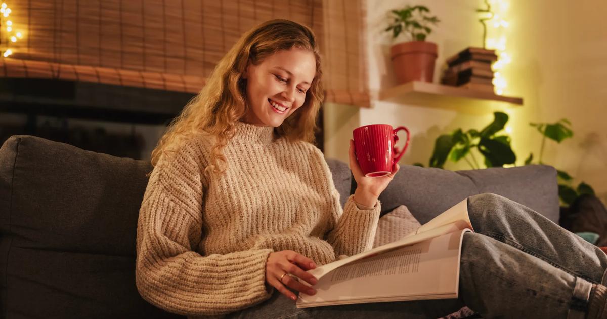 Smiling student reading a book on a couch at home, holding a red mug in a cozy setting.