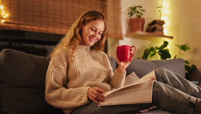Smiling student reading a book on a couch at home, holding a red mug in a cozy setting.