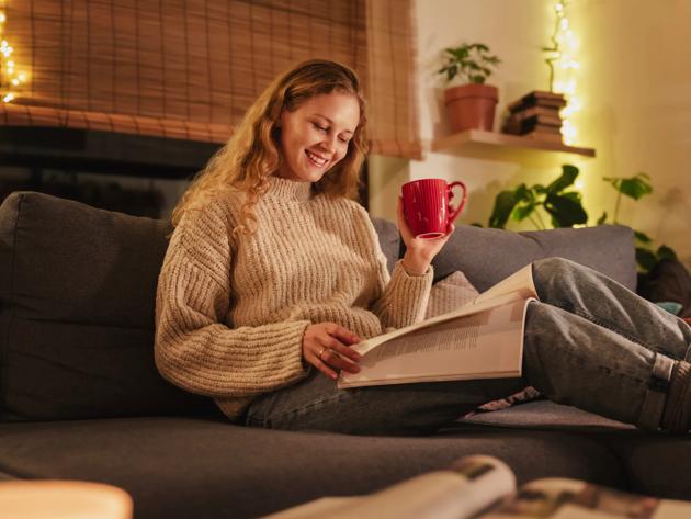 Young pretty curly young blonde woman sitting on sofa at home reading magazine, cozy Christmas holidays