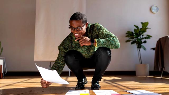 A man kneels as he organizes papers on the floor. 