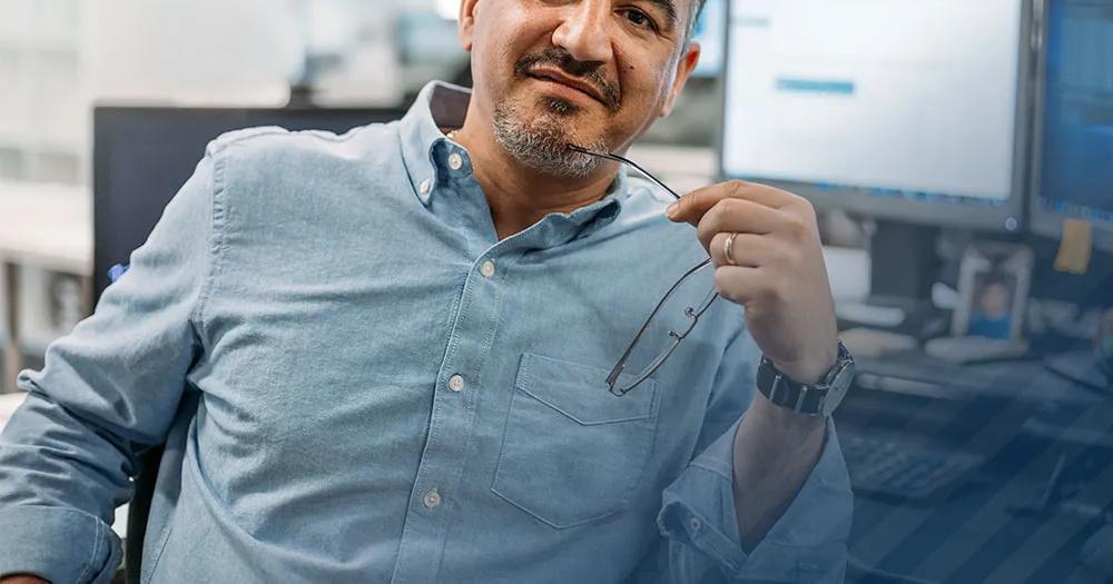 Man sitting at a desk in an office, holding his glasses and looking at the camera.