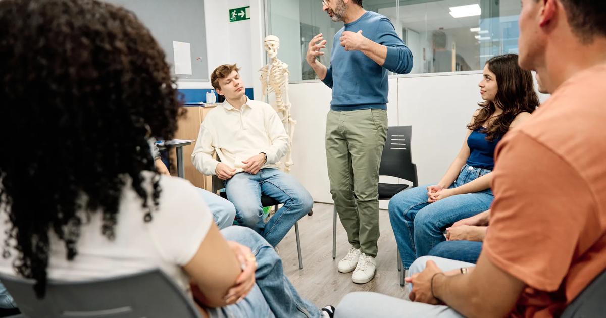 An instructor stands in a circle of students while teaching with a human skeletal model behind him. 