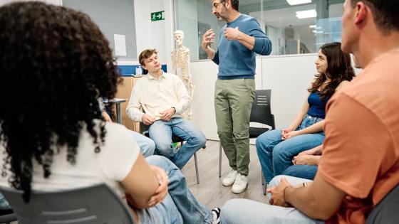 An instructor stands in a circle of students while teaching with a human skeletal model behind him. 