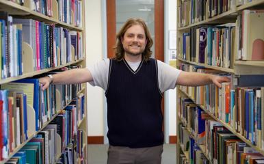 Caleb poses between bookshelves in the library where he worked while earning his degree.