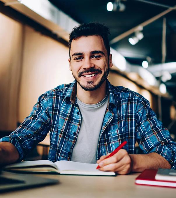 Smiling student sits at computer desk with a laptop and paper notebook