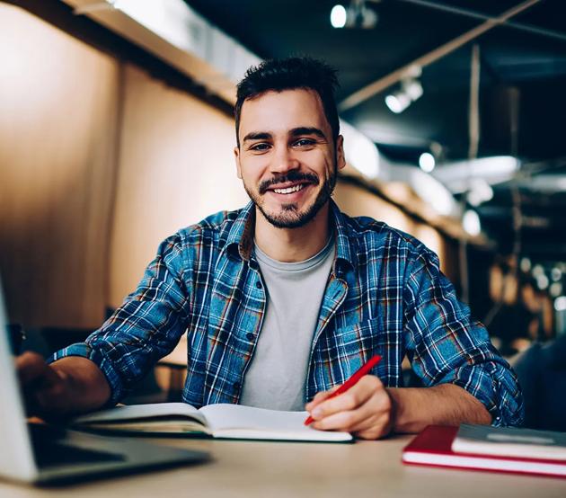 A smiling student seated at a table with a laptop reviews his checklist.