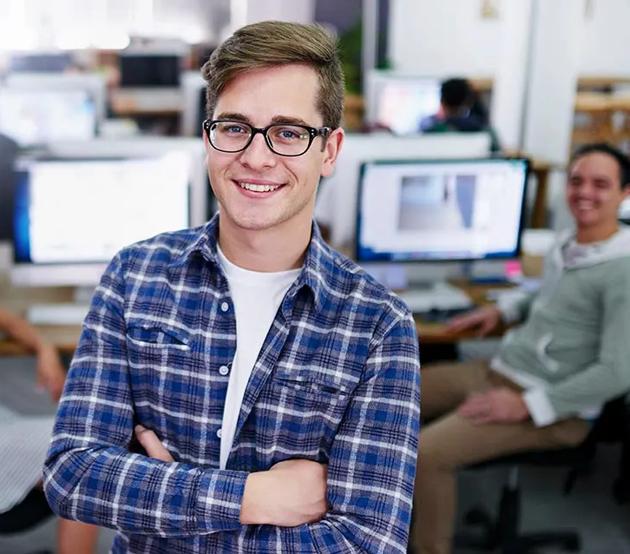 Young college student stands in an airy computer lab. His arms are folded and he is smiling at the camera.