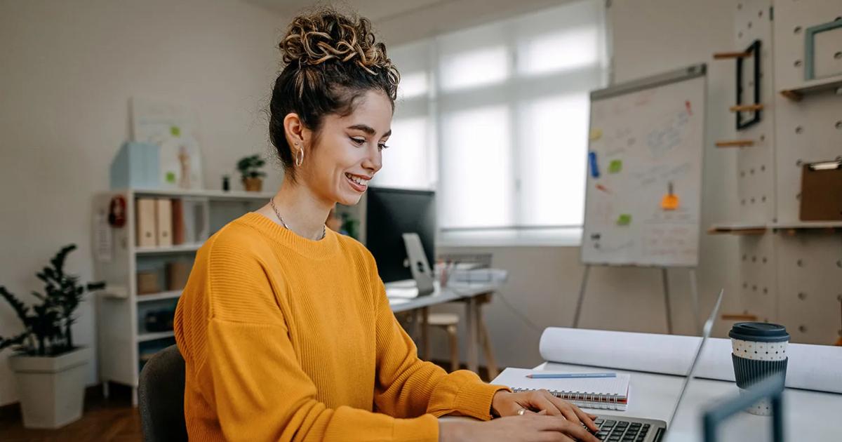 Woman in a yellow sweater smiling while typing on a laptop at a desk in a bright home office.