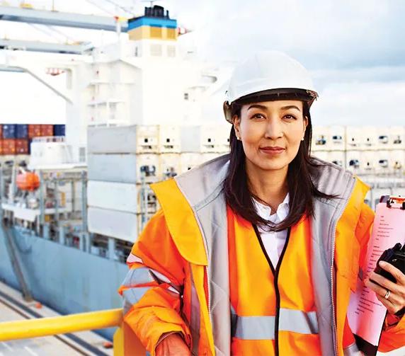 A woman in a hard hat and orange safety jacket stands on a balcony overlooking a cargo port where containers are being unloaded from a shipping vessel.