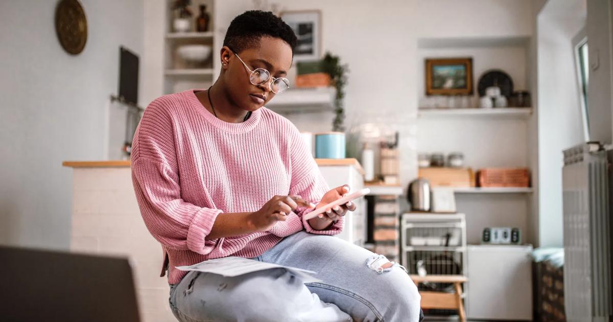A woman in a pink sweater sits in her kitchen with papers on her lap and looks down at her cell phone.