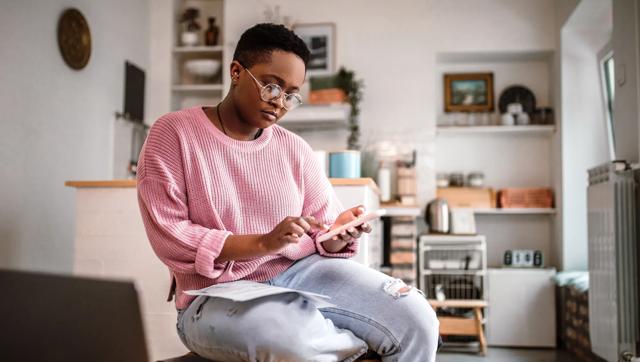 A woman in a pink sweater sits in her kitchen with papers on her lap and looks down at her cell phone.