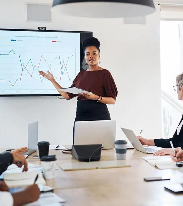 A data analytics professional stands by a chart and presents information to business leaders in a bright conference room.