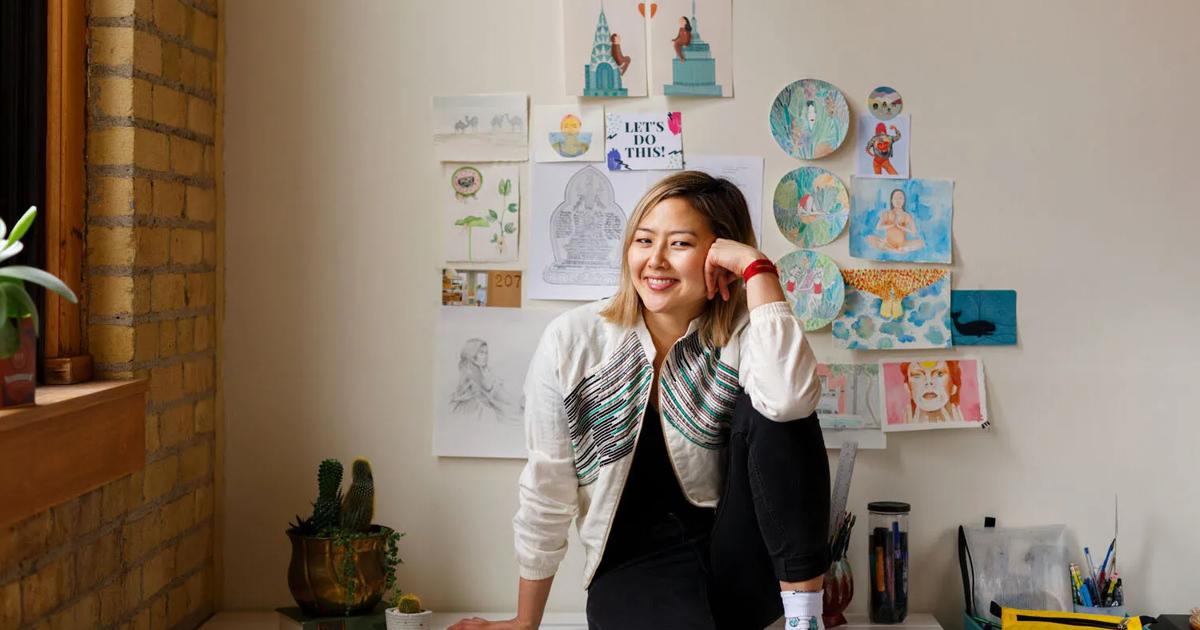 Smiling student sitting on a desk in a creative workspace with artwork displayed on the wall behind her.