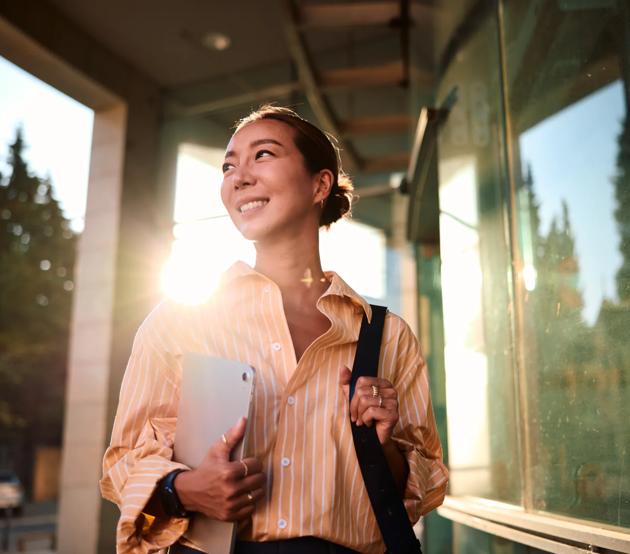 A woman walks past large windows in front of an office building, carrying a laptop in one arm and a portfolio bag over one shoulder.