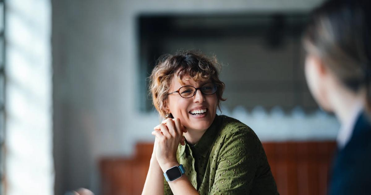 A woman sitting at a table smiles at another person just out of frame. 