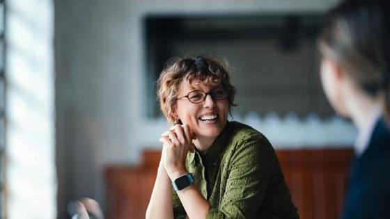 A woman sitting at a table smiles at another person just out of frame. 