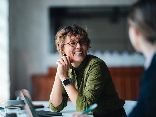 A woman sitting at a table smiles at another person just out of frame. 
