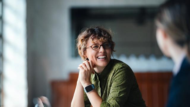 A woman sitting at a table smiles at another person just out of frame. 