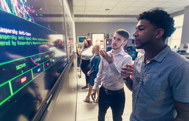 Computer professionals meet in front of a large display screen.