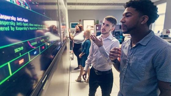 Computer professionals meet in front of a large display screen.