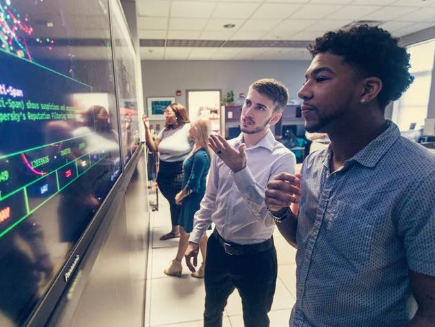Computer professionals meet in front of a large display screen.