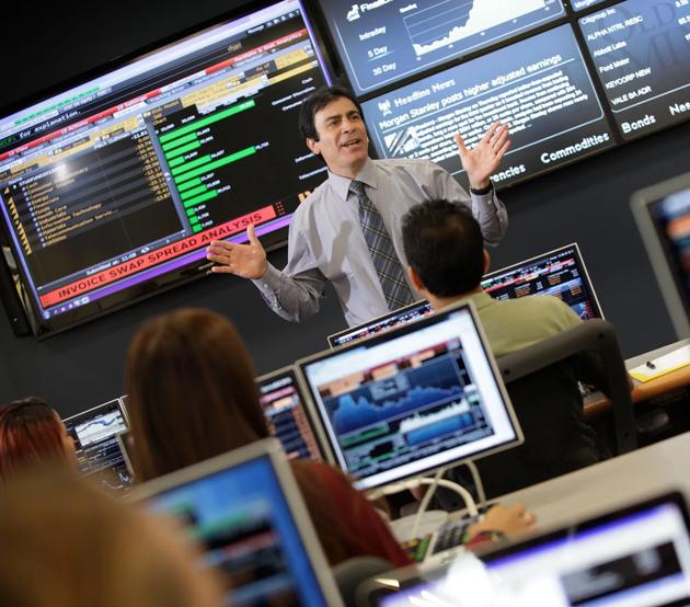 A professor teaches class in front of large digital displays of stock trading data.