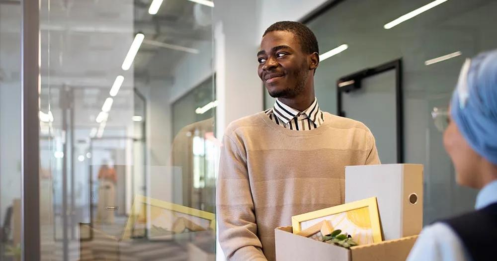 Man holding a box of belongings, smiling in an office hallway.