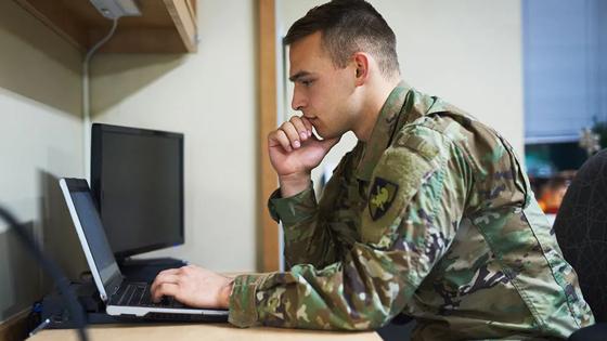 An active duty servicemember prepares for class at a laptop on a desk.