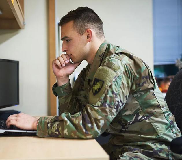 An active duty servicemember prepares for class at a laptop on a desk.