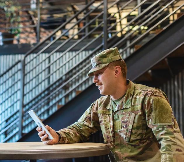A seated active duty military service man looks at a handheld tablet device.