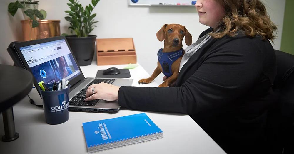 Woman working on a laptop at a desk with a small dog beside her.