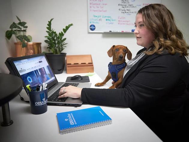 Woman works on her computer with a little dog nearby as she finishes college online