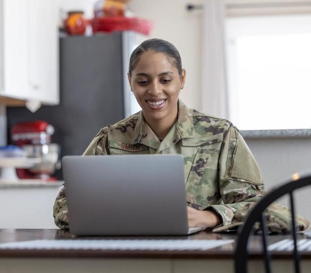 A female military service member logs into class using a laptop on a table at home.