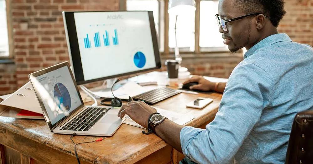 A male African-American college student is studying economic charts on his computer while seated at a wooden desk.