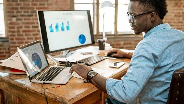 A male African-American college student is studying economic charts on his computer while seated at a wooden desk.