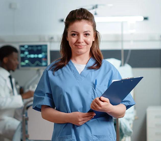 A nurse holding a medical chart stands in a patient's room and smiles with confidence.