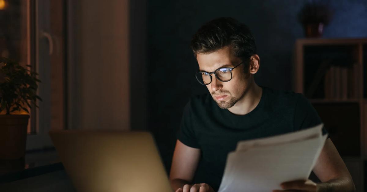 Male student reviewing papers while working on a laptop at night.