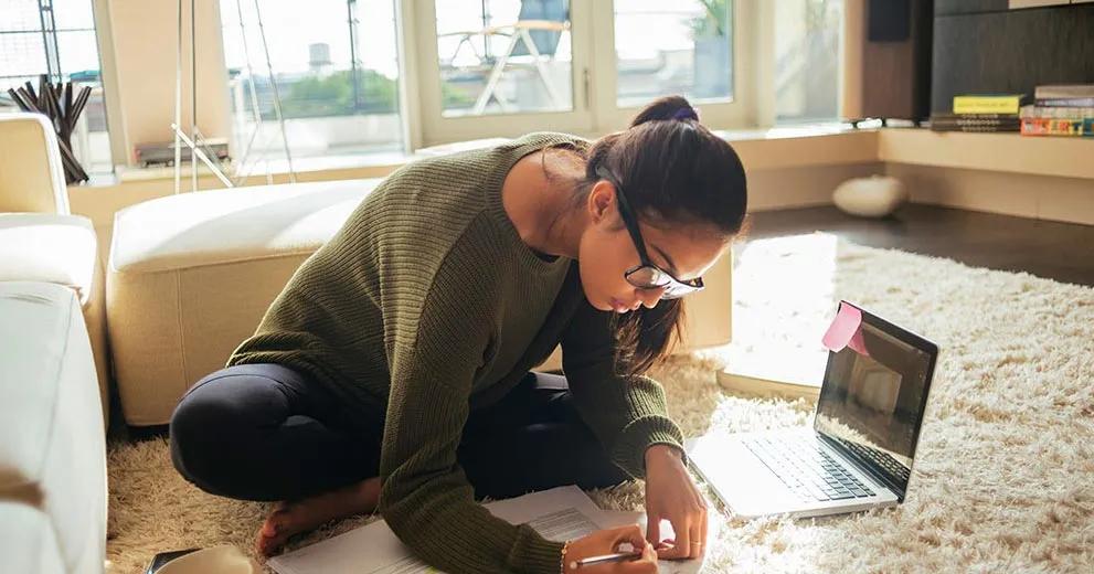 Woman sitting on the floor at home with a laptop and papers beside a cup of coffee.