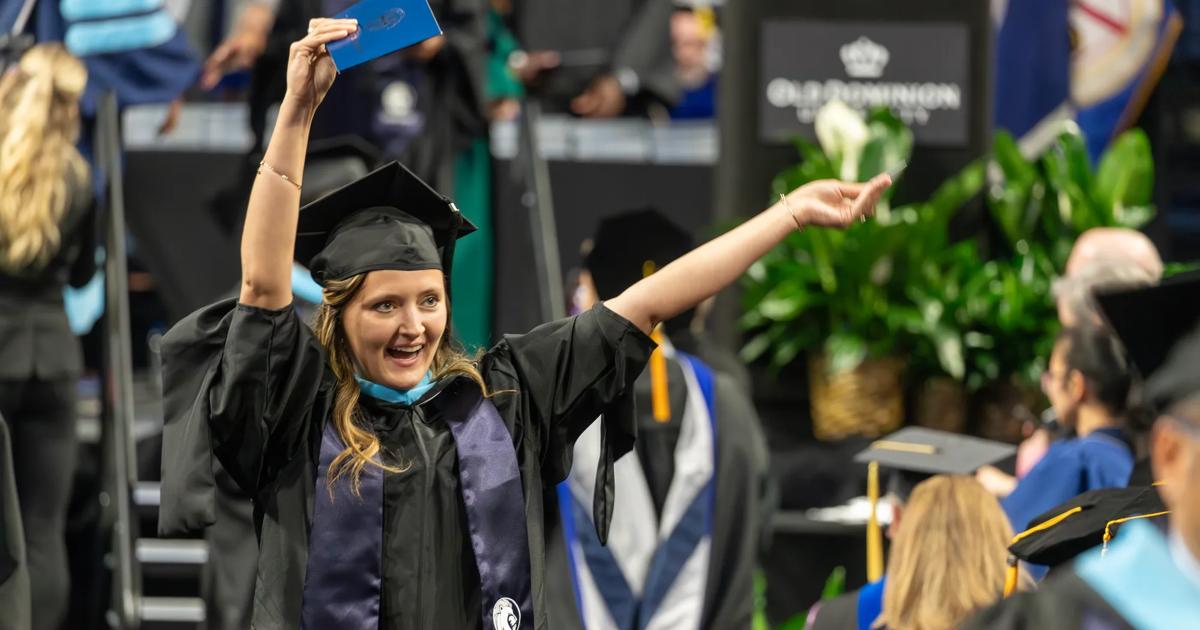 A graduating ODUGlobal student wearing graduation gown and cap waves to family members as she walks in the commencement ceremony.
