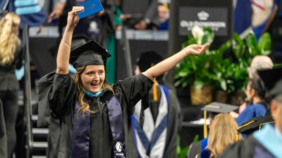 A graduating ODUGlobal student wearing graduation gown and cap waves to family members as she walks in the commencement ceremony.