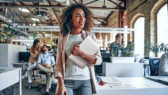 Woman stands in a professional workspace holding a laptop and smiling