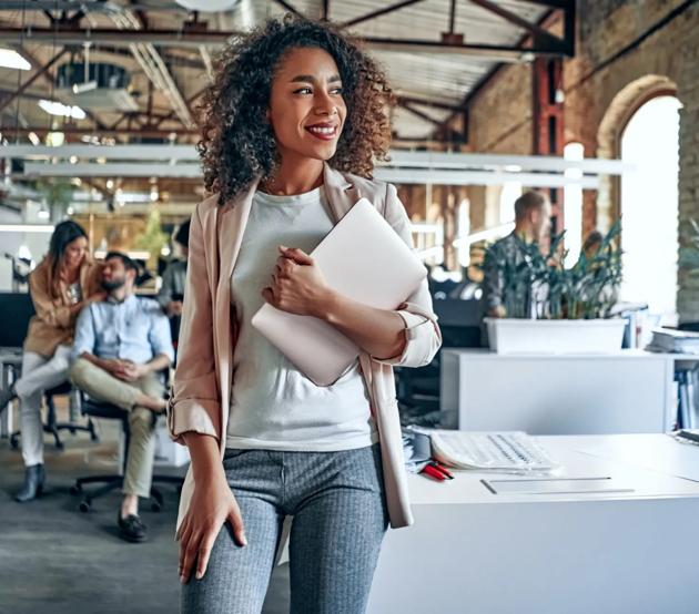Woman stands in a professional workspace holding a laptop and smiling