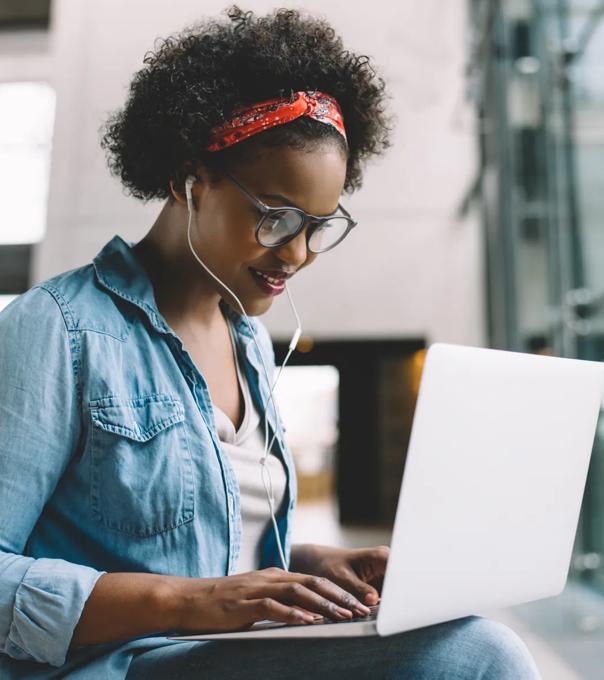 A young woman types on a laptop in a brightly lit room while wearing ear buds.