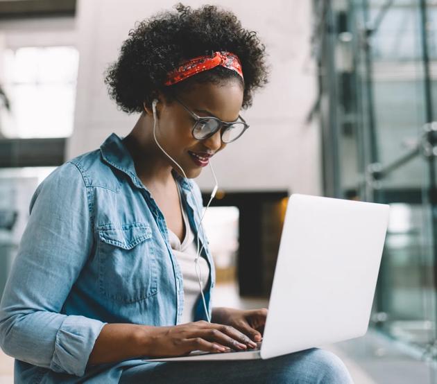 A young woman types on a laptop in a brightly lit room while wearing ear buds.