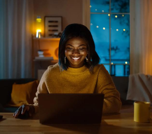 Portrait of Beautiful Smiling Black Girl Working on a Laptop while Sitting at Her Desk at Home. In the Evening Creative Woman Works on a Computer In Her Cozy Living Room. 