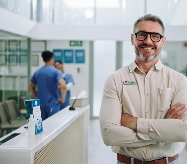 Mature, Medical and Health Professions Education (MHPE) clinician standing with arms crossed and a confident smile in the clinic lobby.